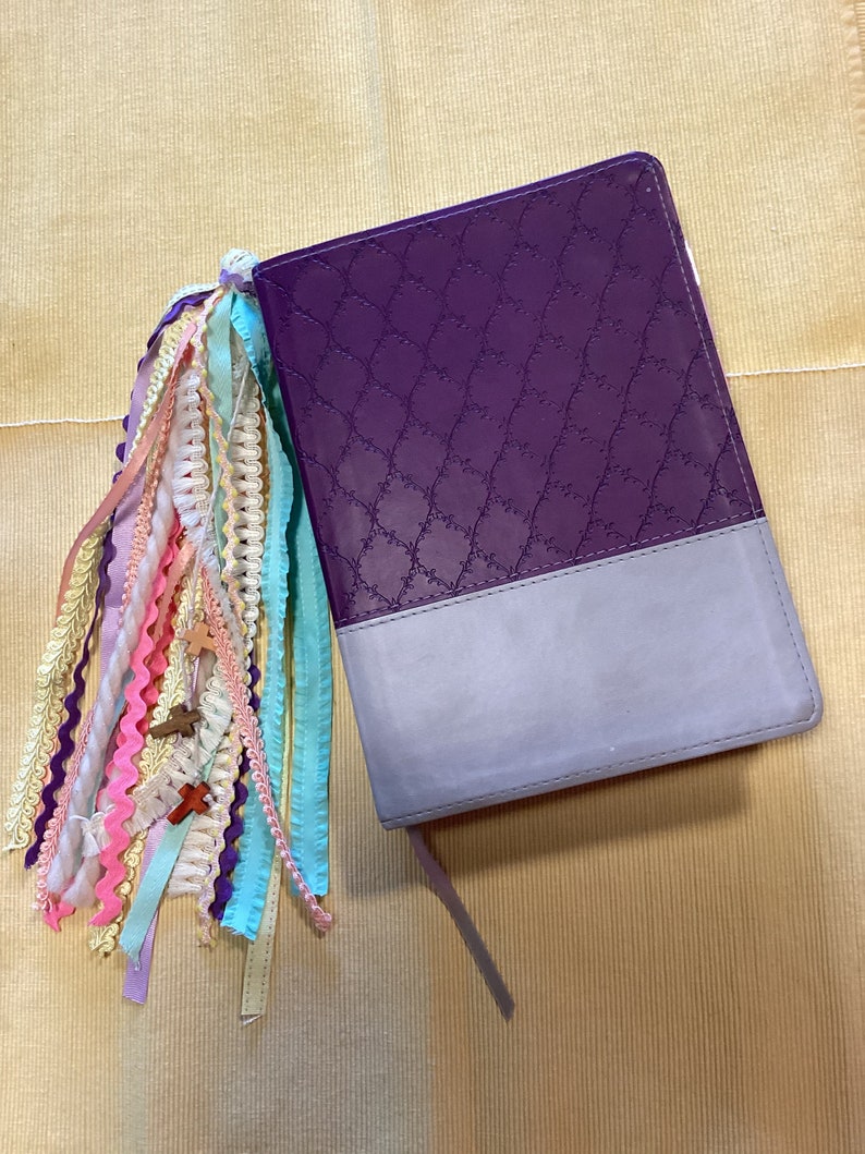 May include: A purple and grey journal with a decorative embossed pattern on the cover. The journal has a long tassel made of colourful ribbons attached to the spine.