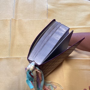 May include: A purple journal with a decorative pattern and a long, colourful ribbon bookmark. The journal is closed and lying on a yellow fabric surface.