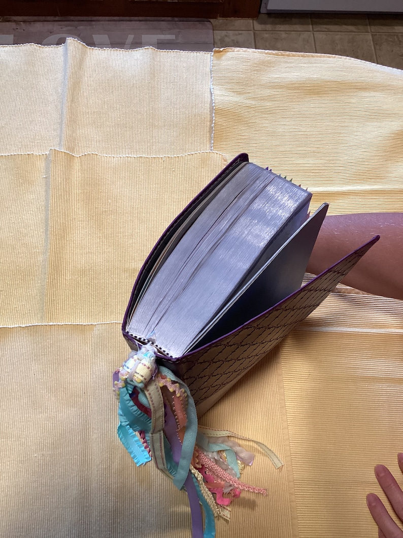 May include: A purple journal with a decorative pattern and a long, colourful ribbon bookmark. The journal is closed and lying on a yellow fabric surface.