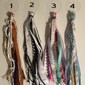 May include: Four different types of colourful ribbon tassels, each with a number tag. The tassels are made of various textures and patterns, including lace, satin, and glitter.