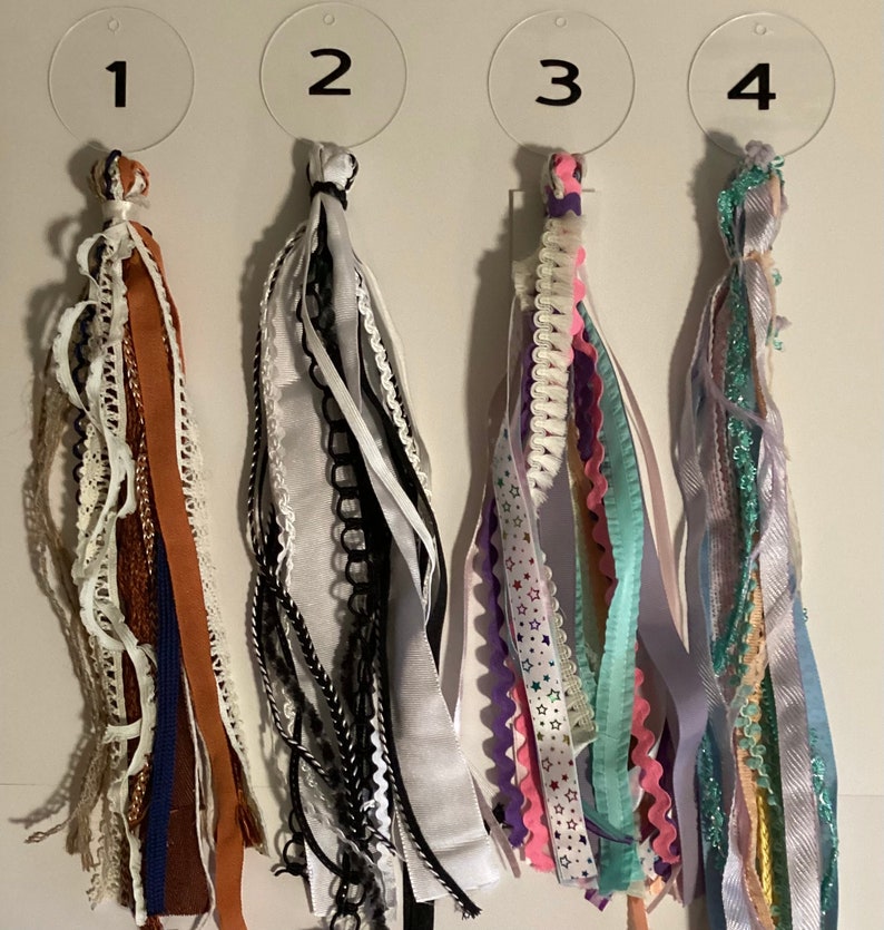 May include: Four different types of colourful ribbon tassels, each with a number tag. The tassels are made of various textures and patterns, including lace, satin, and glitter.