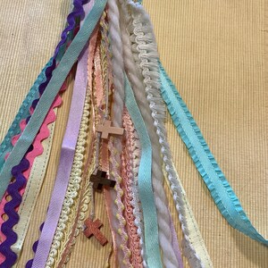 May include: A colourful tassel bookmark with multiple ribbons and a wooden cross charm. The bookmark is made of various textures and colours, including pink, purple, blue, white, and yellow.