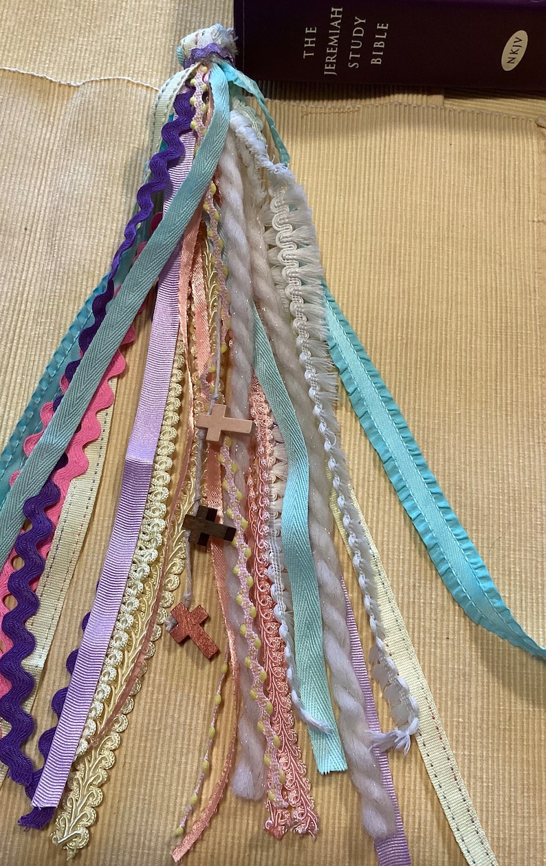 May include: A colourful tassel bookmark with multiple ribbons and a wooden cross charm. The bookmark is made of various textures and colours, including pink, purple, blue, white, and yellow.