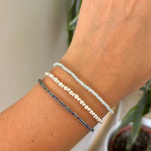 May include: Three beaded bracelets stacked on a wrist. The top bracelet is light blue, the middle bracelet is white with silver beads, and the bottom bracelet is dark blue with iridescent beads.
