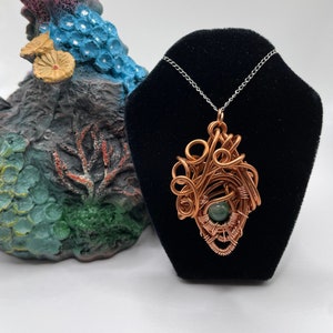 May include: A copper wire wrapped pendant with a green gemstone in the center. The pendant is hanging from a silver chain on a black velvet display stand.