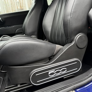 May include: Black leather car seat with white stitching and a grey suede insert. The seat has a black base with the number "500" in white lettering. The car's interior is visible in the background.