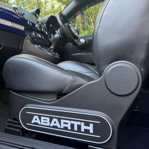 May include: Close-up of a black leather car seat with white stitching. The seat has a black panel with the white "ABARTH" logo. The interior includes a steering wheel and dashboard.
