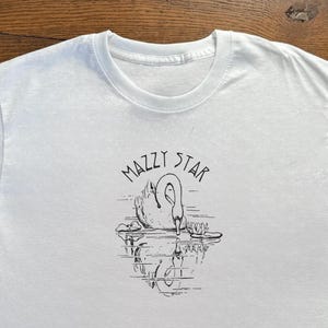 May include: White t-shirt with a black and white swan illustration and the text "MAZZY STAR". The swan is shown in a pond with its reflection. The tee has a crew neck and short sleeves. The shirt is made of a soft material.