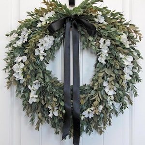 May include: A round wreath composed of green leaves and white flowers, accented with a black satin bow and streamers. The wreath is designed for door or wall decor, offering a natural and elegant aesthetic.