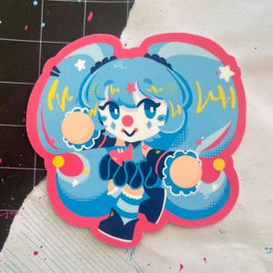 Puede incluir: Una pegatina con una chica payaso de dibujos animados con pelo azul y un contorno rosa. El payaso tiene una nariz roja y sostiene dos objetos redondos. El diseño incluye detalles en amarillo y blanco.