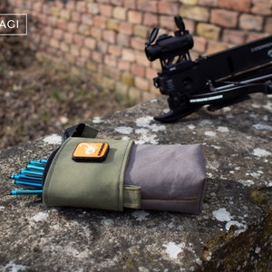 Könnte beinhalten: Ein grüner und grauer Stoffköcher mit blauen Pfeilen und einem kleinen orangenen Patch mit einem Skorpion-Logo. Der Köcher liegt auf einer Steinoberfläche neben einer schwarzen Armbrust mit einem roten Zielfernrohr.
