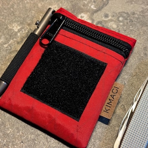 Könnte beinhalten: Eine rote Reißverschlusstasche mit einem schwarzen Klettverschluss-Patch und dem Wort "KIMACI" auf der Seite. Ein silberner Stift und ein silbernes Taschenmesser liegen neben der Tasche.