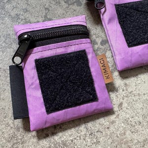 Könnte beinhalten: Zwei kleine, rechteckige, lila Beutel mit schwarzen Reißverschlüssen und schwarzen Klettverschlüssen. Jeder Beutel hat einen schwarzen Riemen und ein kleines braunes Etikett mit "KIMAGI".