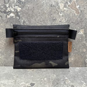 Könnte beinhalten: Schwarz getarnte Reißverschlusstasche mit Klettverschluss und einem kleinen Etikett mit der Aufschrift "KIMACI". Die Tasche hat zwei schwarze Riemen auf jeder Seite. Die Tasche ist aus strapazierfähigem Material gefertigt.