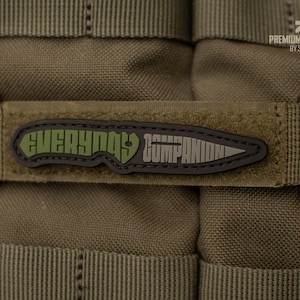 Könnte beinhalten: Nahaufnahme eines olivgrünen Aufnähers mit den Worten "EVERYDAY COMPANION" in Grün und Grau, mit einer Messer-Grafik. Der Aufnäher ist an einem passenden olivgrünen Riemen befestigt, vor einem strukturierten Hintergrund.