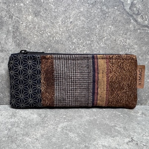 Può includere: Astuccio rettangolare con design patchwork. La custodia presenta un motivo geometrico blu navy, tweed marrone e sezioni a righe in marrone, oro e bordeaux. Una piccola etichetta in pelle recita "KIMACI".