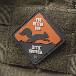 Könnte beinhalten: Ein rautenförmiger Aufnäher mit einem laufenden Eichhörnchen-Silhouette auf orangefarbenem Grund. Der Aufnäher trägt den Text "YOU BETTER RUN" und "LITTLE SQUIRREL". Der Aufnäher ist schwarz und grau und auf einem grünen Stoff befestigt.