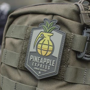 May include: A rectangular patch with a pineapple grenade design, featuring a yellow and green color scheme. The patch is affixed to a green backpack and includes the text "PINEAPPLE EXPRESS AIRMAIL DELIVERY".