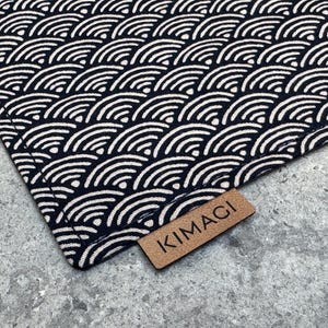 Pode incluir: Close-up de um tecido com um padrão de ondas repetidas em branco sobre um fundo azul marinho. Uma pequena etiqueta retangular de couro castanho com a palavra "KIMACI" está presa ao tecido.