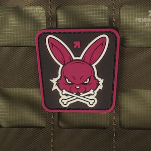 Könnte beinhalten: Ein quadratischer Aufnäher mit einem magentafarbenen Kaninchenkopf und gekreuzten Knochen auf schwarzem Hintergrund. Der Aufnäher hat einen magentafarbenen Rand und befindet sich auf einer grünen, strukturierten Oberfläche. Der Text "PREMIUM PATCHES BY SEVAL" ist sichtbar.