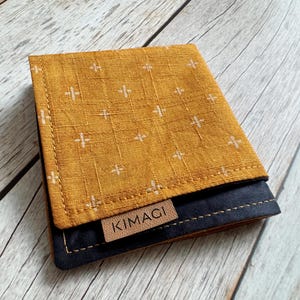 Op de afbeelding: Een kleine, mosterdgele stoffen portemonnee met een wit kruispatroon. De portemonnee heeft een zwarte leren klep met het woord "KIMAGI" in bruin gegraveerd.