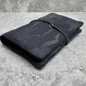 Puede incluir: Un objeto rectangular plegado de tela de camuflaje negra, asegurado con una correa oscura. La tela tiene un sutil patrón de verde oscuro y gris. El objeto está sobre una superficie gris texturizada.