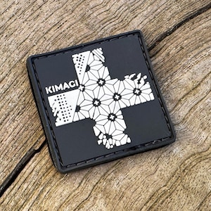 Könnte beinhalten: Schwarzer quadratischer Aufnäher mit einem weißen Kreuzdesign mit geometrischen Blumenmustern. Das Wort "KIMAGI" ist weiß aufgedruckt. Der Aufnäher liegt auf einer strukturierten Holzoberfläche.