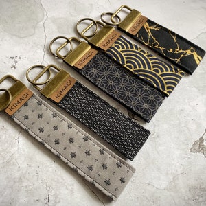 Könnte beinhalten: Fünf Schlüsselanhänger mit gold- und schwarz gemustertem Stoff und dem Wort "KIMAGI" auf einem braunen Lederanhänger.