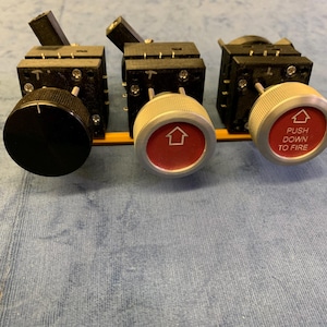 May include: Three black and silver control switches with knobs. The first knob is black, the second and third are silver with red centers. The second knob has an upward-pointing arrow, and the third says "PUSH DOWN TO FIRE".
