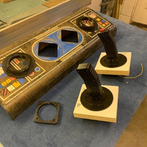 May include: An arcade game control panel with two black joysticks with red buttons, and a separate black square ring. The panel has blue and yellow accents and two square holes.