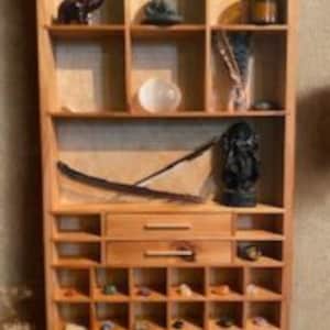 May include: A light-colored wooden display shelf featuring multiple square compartments, two central drawers with horizontal handles, and various decorative items including small stones, figurines, a white sphere, and a dark feather.