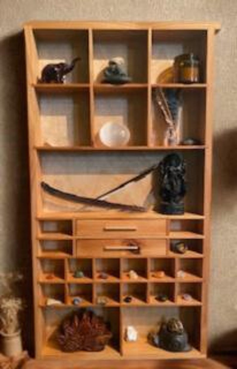 May include: A light-colored wooden display shelf featuring multiple square compartments, two central drawers with horizontal handles, and various decorative items including small stones, figurines, a white sphere, and a dark feather.