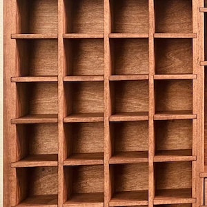 Puede incluir: Una vitrina rectangular de madera que presenta una cuadr&iacute;cula de m&uacute;ltiples compartimentos cuadrados. La unidad est&aacute; construida con madera de tono marr&oacute;n medio con una veta visible, organizada en cuatro columnas verticales y siete filas horizontales.