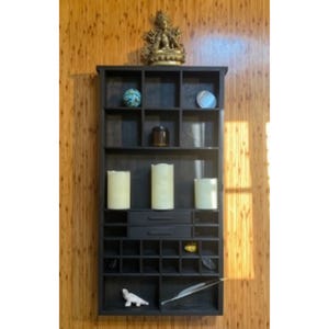 May include: A dark brown wooden wall-mounted organizer featuring multiple square compartments, two central drawers, and various decorative items including three pillar candles, a small statue, a blue sphere, and a white feather quill.