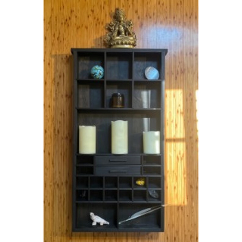 May include: A dark brown wooden wall-mounted organizer featuring multiple square compartments, two central drawers, and various decorative items including three pillar candles, a small statue, a blue sphere, and a white feather quill.
