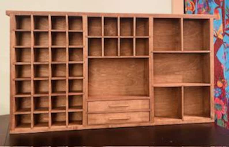 Puede incluir: Un estante organizador de madera marr&oacute;n que presenta una cuadr&iacute;cula de veinticuatro compartimentos cuadrados peque&ntilde;os a la izquierda, una secci&oacute;n con ocho cub&iacute;culos m&aacute;s peque&ntilde;os sobre un espacio abierto m&aacute;s grande y dos cajones horizontales en el centro, y tres estantes abiertos grandes a la derecha.
