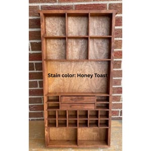 May include: A wooden wall-mounted display shelf with a honey toast stain finish. The unit features three large upper compartments, two central drawers with horizontal handles, and a grid of smaller square cubbies at the bottom for storage.