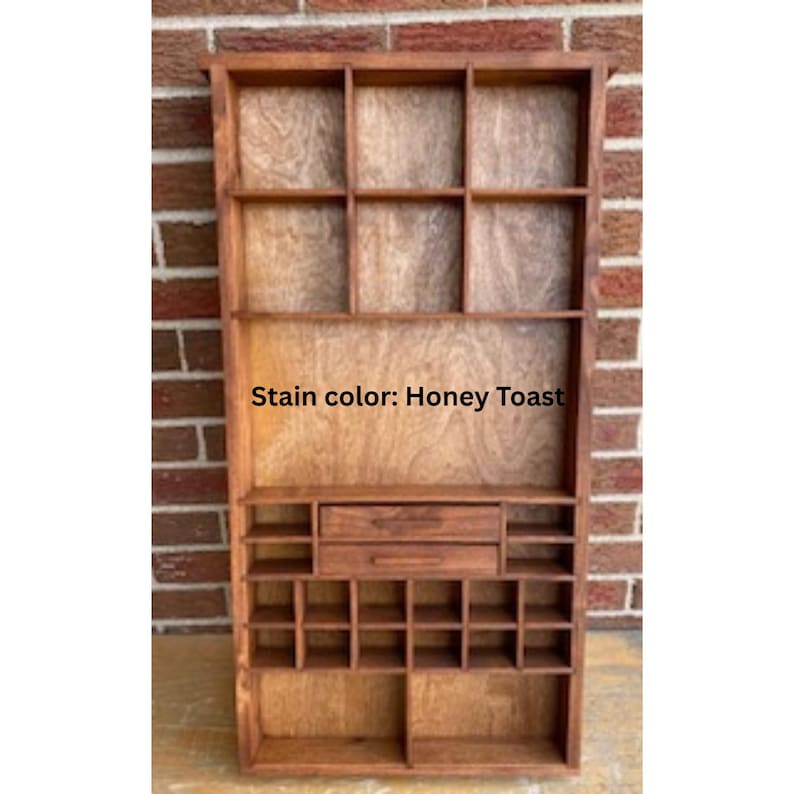 May include: A wooden wall-mounted display shelf with a honey toast stain finish. The unit features three large upper compartments, two central drawers with horizontal handles, and a grid of smaller square cubbies at the bottom for storage.