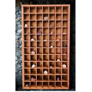 May include: A wooden display shelf with numerous small square compartments. The shelf is filled with various rocks and crystals in different colors and shapes. The wood has a warm, natural tone, and the shelf is mounted against a textured stone wall.