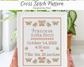 May include: A framed cross-stitch pattern with the text "Francesa Sofia Ricci" and birth details. The design includes butterflies in shades of green, peach, and brown. The frame is white, and the text reads "October 14, 2022, 4:30 am, 7lbs. 9oz. 20 inches."