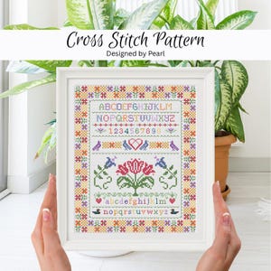 May include: A framed cross stitch pattern with floral and bird motifs. The design includes the alphabet, numbers, and decorative elements in a colorful, folk art style. The text "Cross Stitch Pattern" and "Designed by Pearl" are at the top.