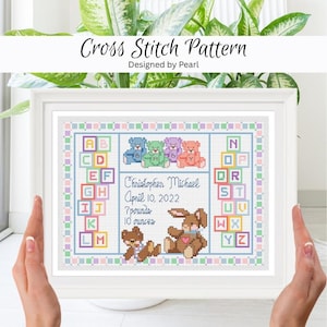 May include: A framed cross-stitch pattern with a baby theme. The design includes colorful teddy bears, a bunny, and the text "Christopher Michael, April 10, 2022, 3.2 kg, 283 grams." The frame is white, and the background is a soft pastel color.
