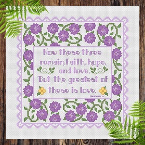 Peut inclure: Un motif de point de croix sur tissu blanc avec une bordure florale violette et le texte : Now these three remain, faith, hope, and love. But the greatest of these is love. Corinthians 13. Le design inclut des vignes vertes et des fleurs jaunes.