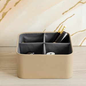 Op de afbeelding: Een beige, vierkante opbergdoos met een stoffen voering en vier vakken. De doos is gevuld met make-upkwasten en kleine containers.
