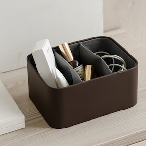 Könnte beinhalten: Eine braune lederartige Box mit drei Fächern. Die Box enthält eine weiße Box, eine goldene Flasche und eine Rolle weißen Kordels. Die Box steht auf einer hellbraunen Holzoberfläche.