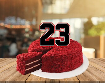 May include: A red velvet cake with white frosting and a number 23 on top. The cake is on a white plate and a wooden table.