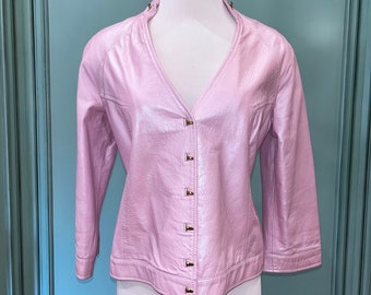 Puede incluir: Una chaqueta de cuero rosa con cuello en V y cierre de botones. La chaqueta tiene un acabado brillante y botones dorados.