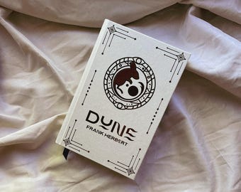 Może przedstawiać: Książka w twardej oprawie zatytułowana "DUNE" autorstwa Franka Herberta. Książka ma białą okładkę z okrągłym emblematem przedstawiającym brązowe stworzenie i geometryczne wzory. Tytuł jest w stylizowanej czcionce i spoczywa na pomarszczonej, jasnej tkaninie.