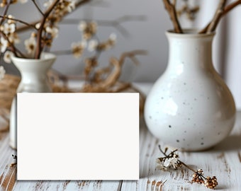 May include: Two white ceramic vases with branches of white flowers. A blank white card sits in front of the vases on a white wooden surface. The background is blurred, with a neutral color palette.