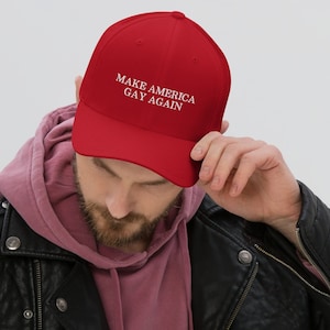 May include: A red baseball cap with the text "MAKE AMERICA GAY AGAIN" embroidered in white. The cap is worn by a person wearing a black leather jacket and a pink hoodie.